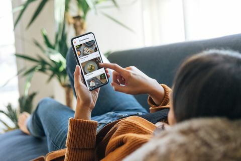 over skulderutsikten til en ung kvinne som velger mat fra menyen i mobilappen mens hun ligger på sofaen hjemme