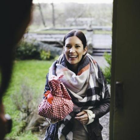 en kvinne smiler mens hun står på dørene og besøker en venn hun er pakket inn i varme klær og holder en gave innpakket i en rød og hvit rutet klut