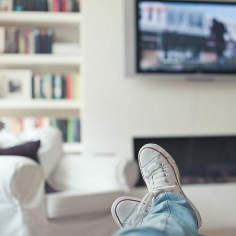 Bilde av jeans og joggesko, føttene opp på sofaen, med TV, peis, sofa og bokhylle i bakgrunnen.